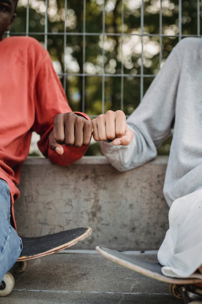 Two teenagers sharing a fist bump while sitting with skateboards outdoors.